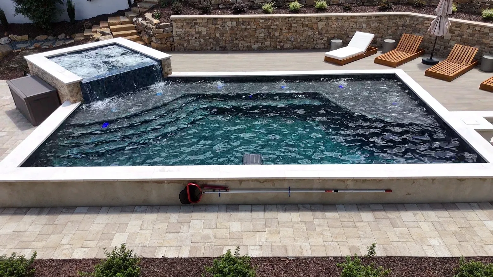 Angled drone view of a raised spa and pool set into a terraced backyard with stone retaining walls