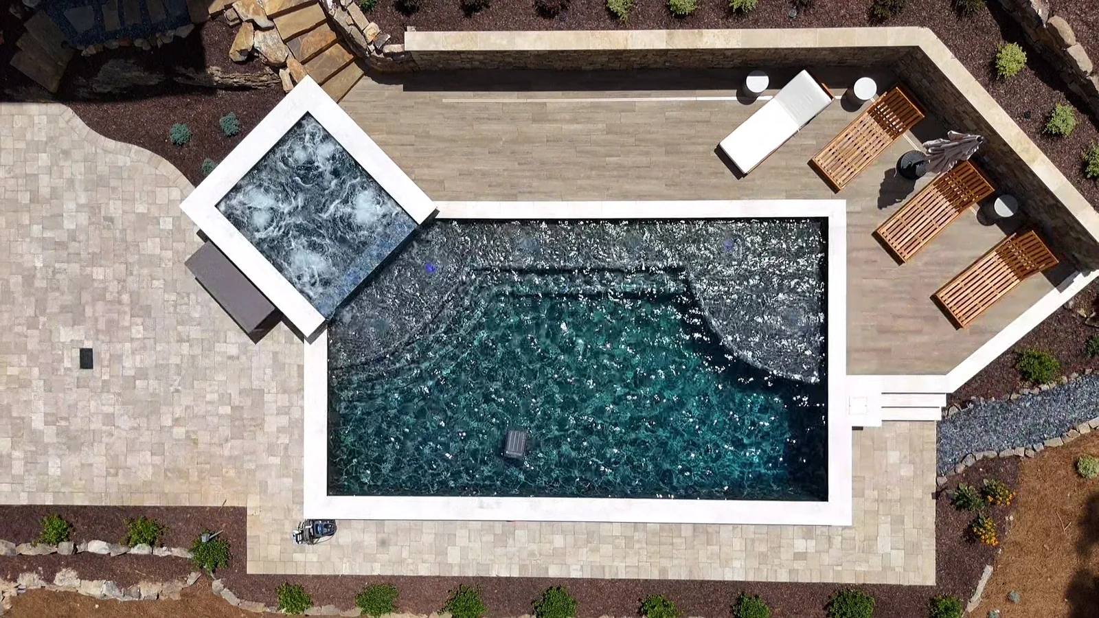 Straight-down drone view of a raised spa spilling into a dark-tile pool with a full paver patio and loungers