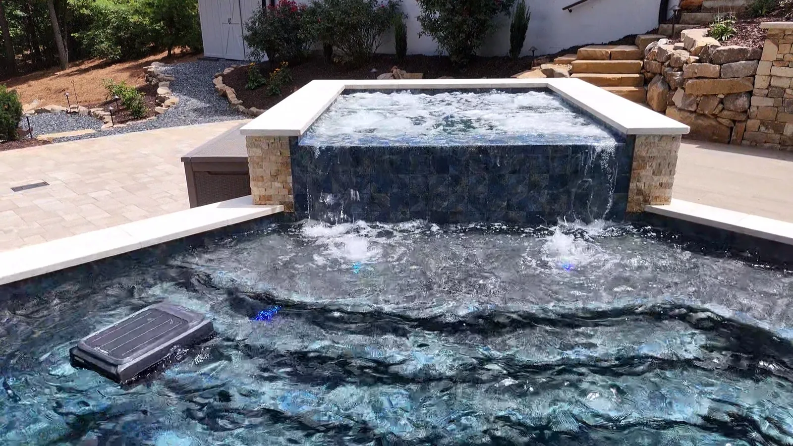 Raised spa sheeting water over a dark tiled wall into the pool below, framed by a stacked-stone retaining wall