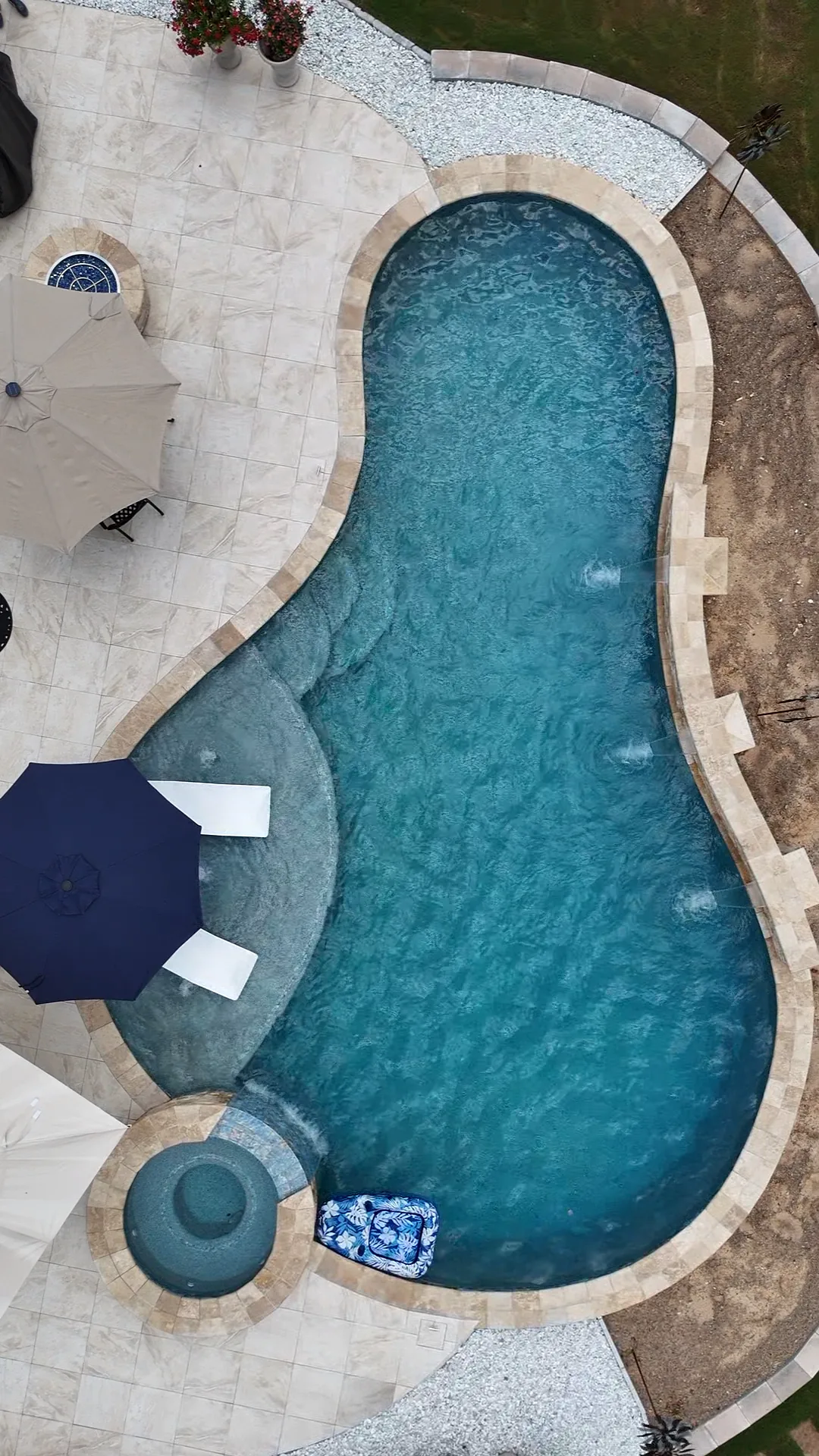 Straight-down aerial view of the completed Curtis pool, round spa, pale stone patio, umbrellas, and tanning ledge