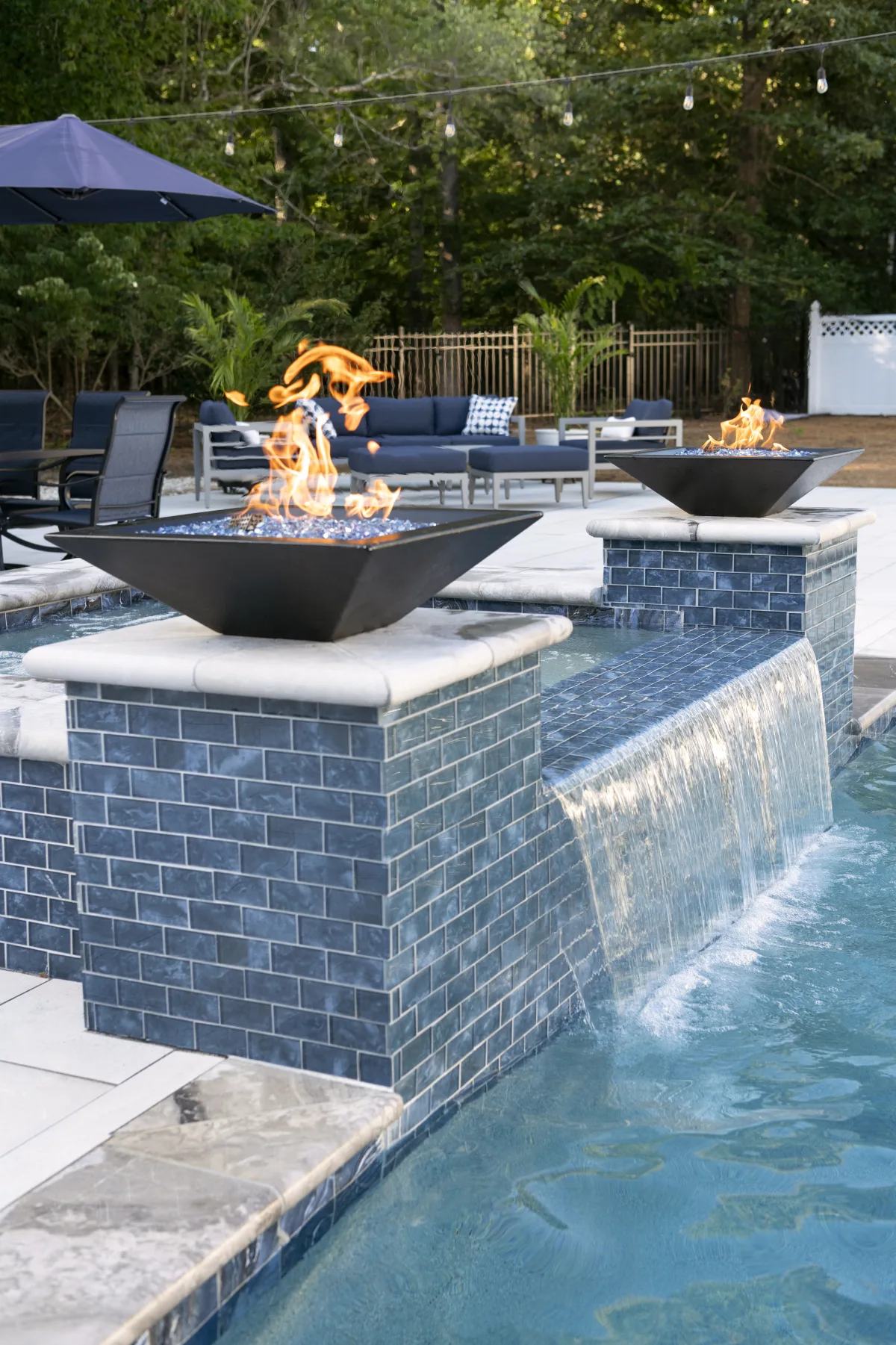 Two fire bowls burning on blue-tiled columns above a waterfall spilling into a RutherfordMade pool